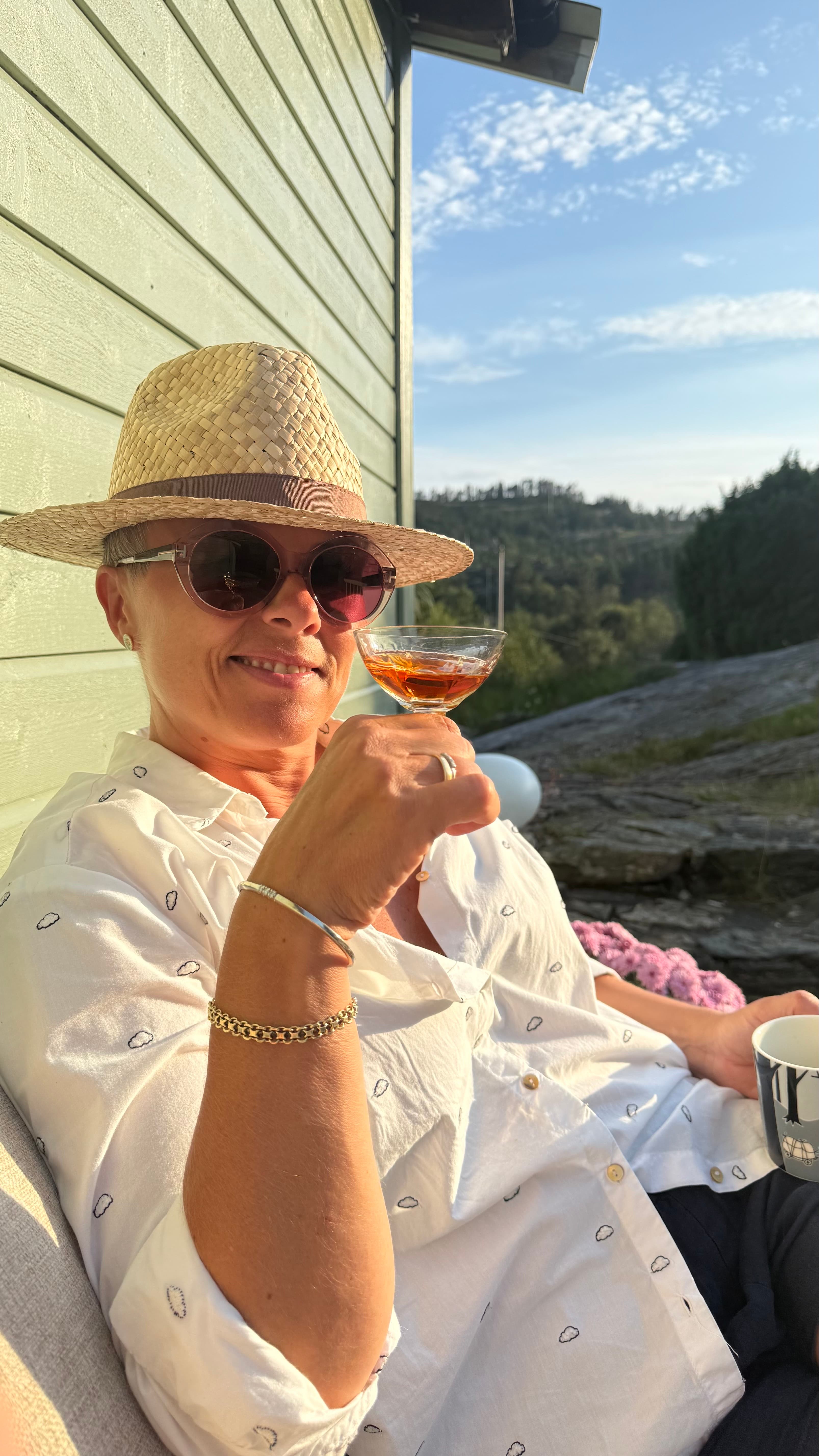 Johanne sitter ved hytteveggen i kveldssol med hatt og et glass i hÄnden.